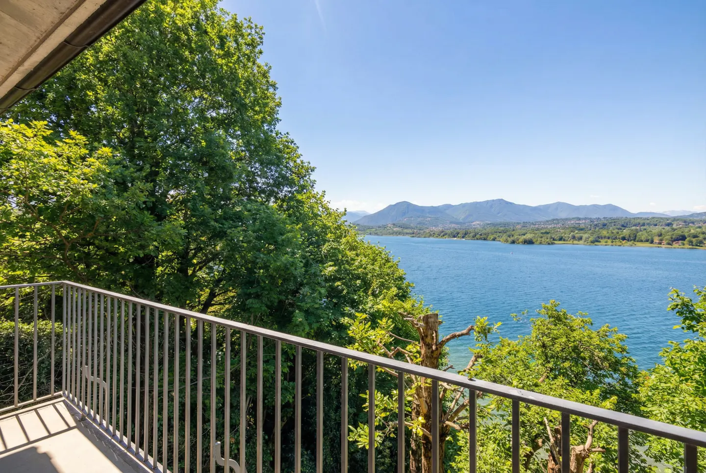 Balcony view of a blue lake surrounded by green trees and mountains under a clear blue sky. Metal railing in the foreground.