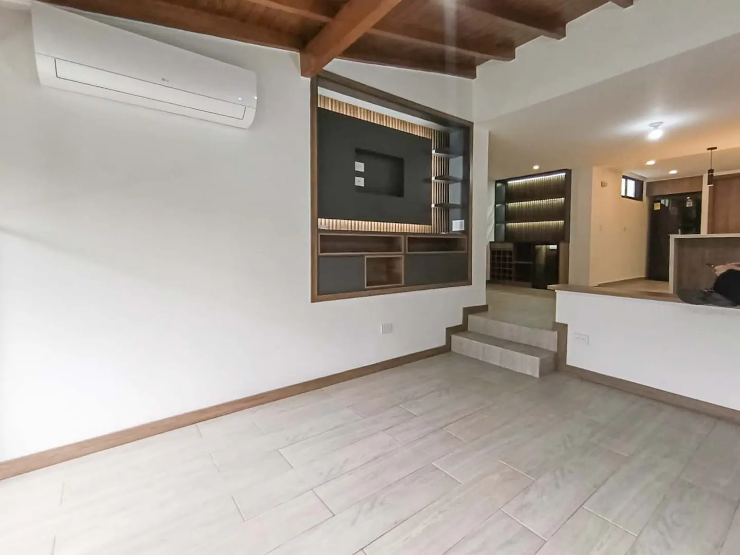 Bright, modern living room with white walls, light wood floors, and a wooden ceiling. A built-in entertainment center and steps lead to an open kitchen.