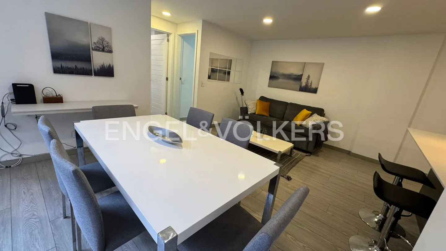 A modern apartment interior with a white dining table, gray chairs, and a gray sofa with yellow pillows. Artwork adorns the white walls.