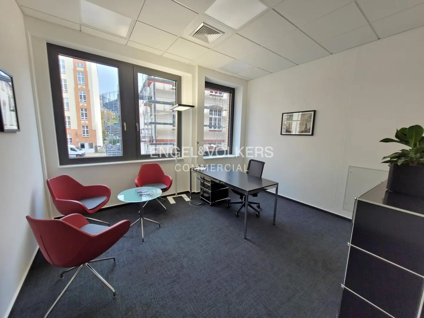 Office space with black desk and chair, red chairs around a glass table, and large windows.