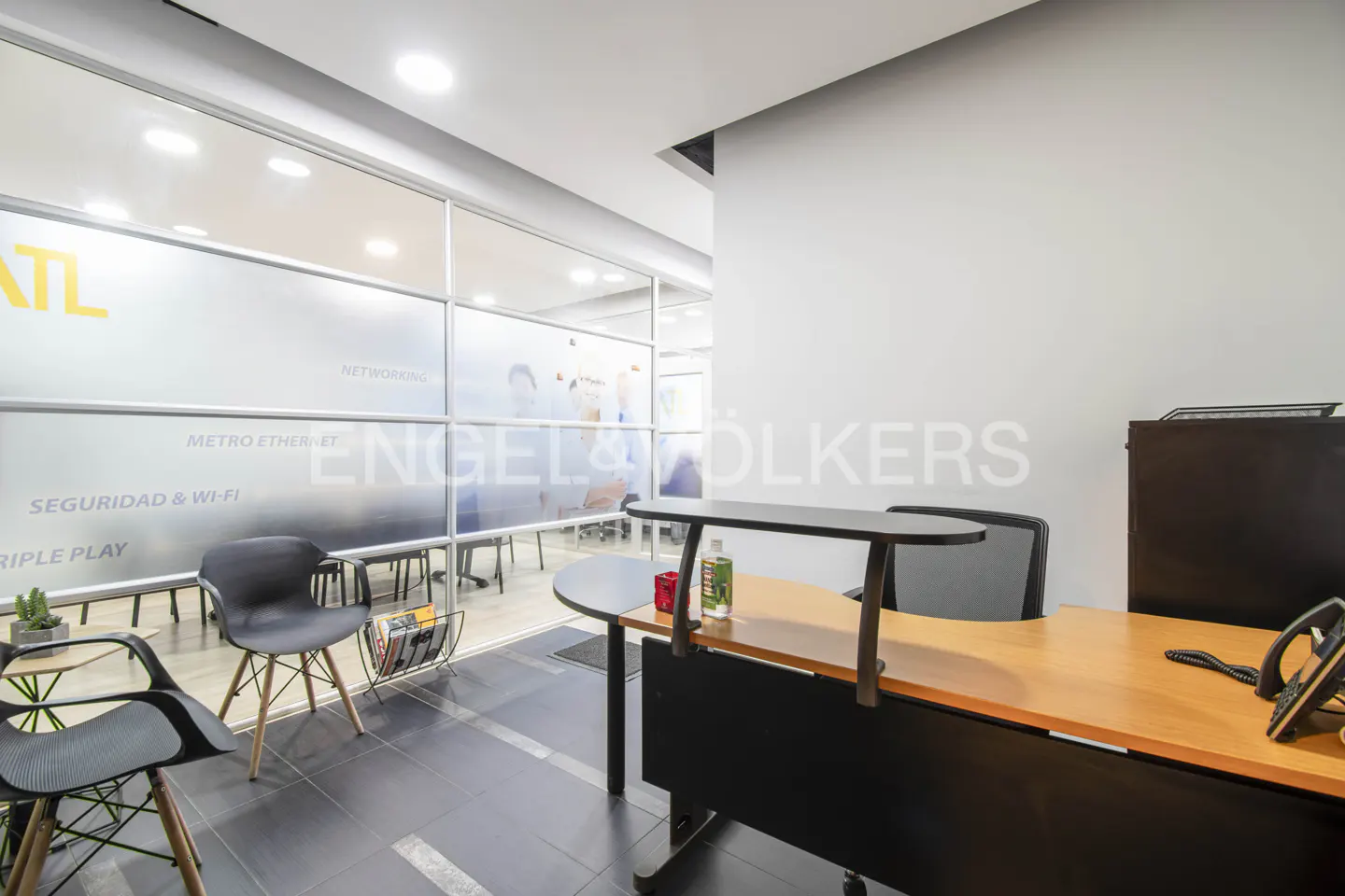 Modern office reception area with a wooden desk, black chairs, and a glass-walled meeting room in the background.