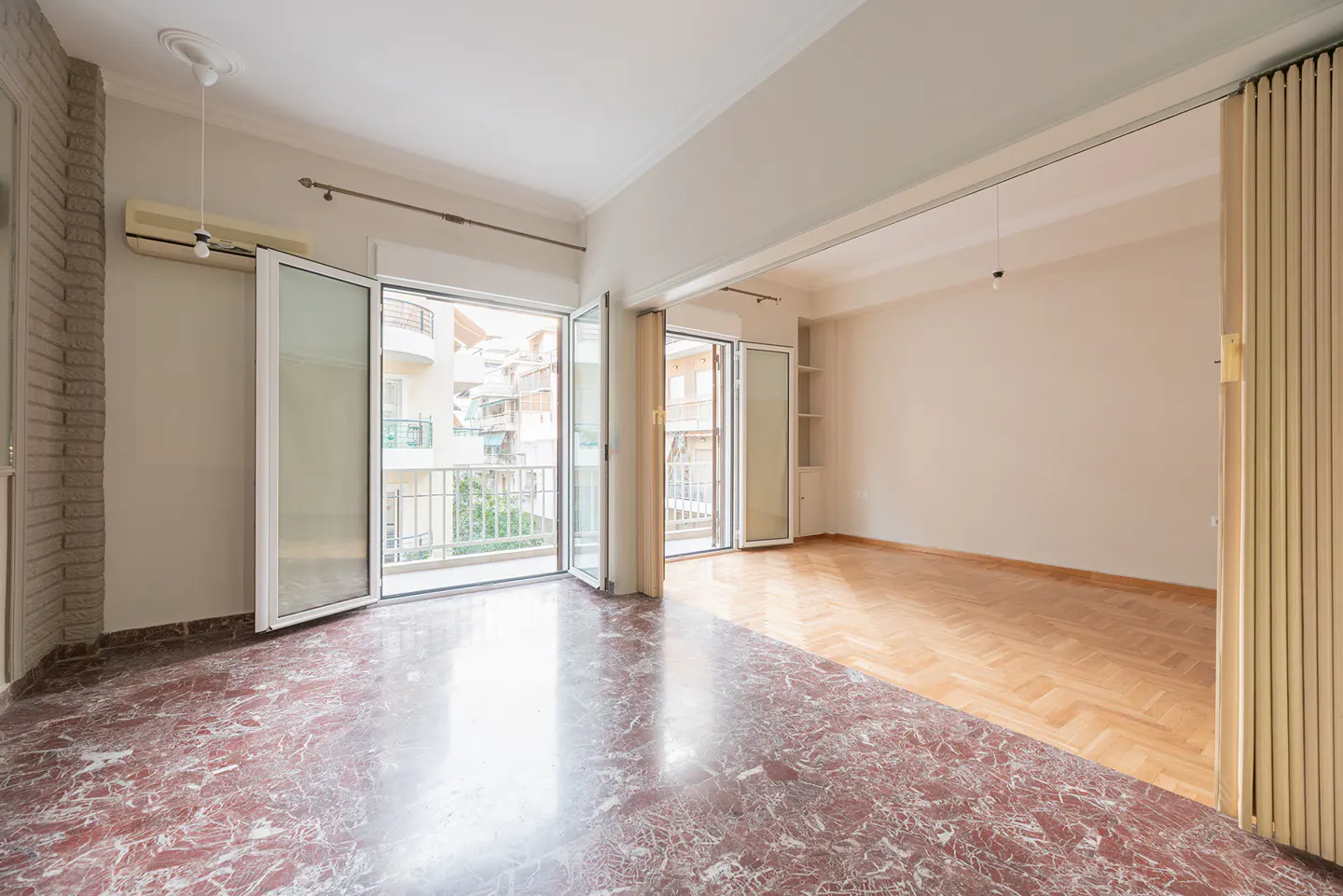 Bright, empty room with red marble and wood floors. Balconies are visible through open glass doors. Cream walls and white trim.