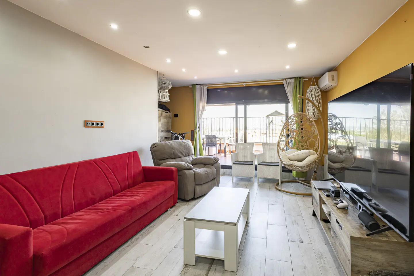 Modern living room with a bright red sofa, grey recliner, white coffee table, hanging egg chair, and large TV.