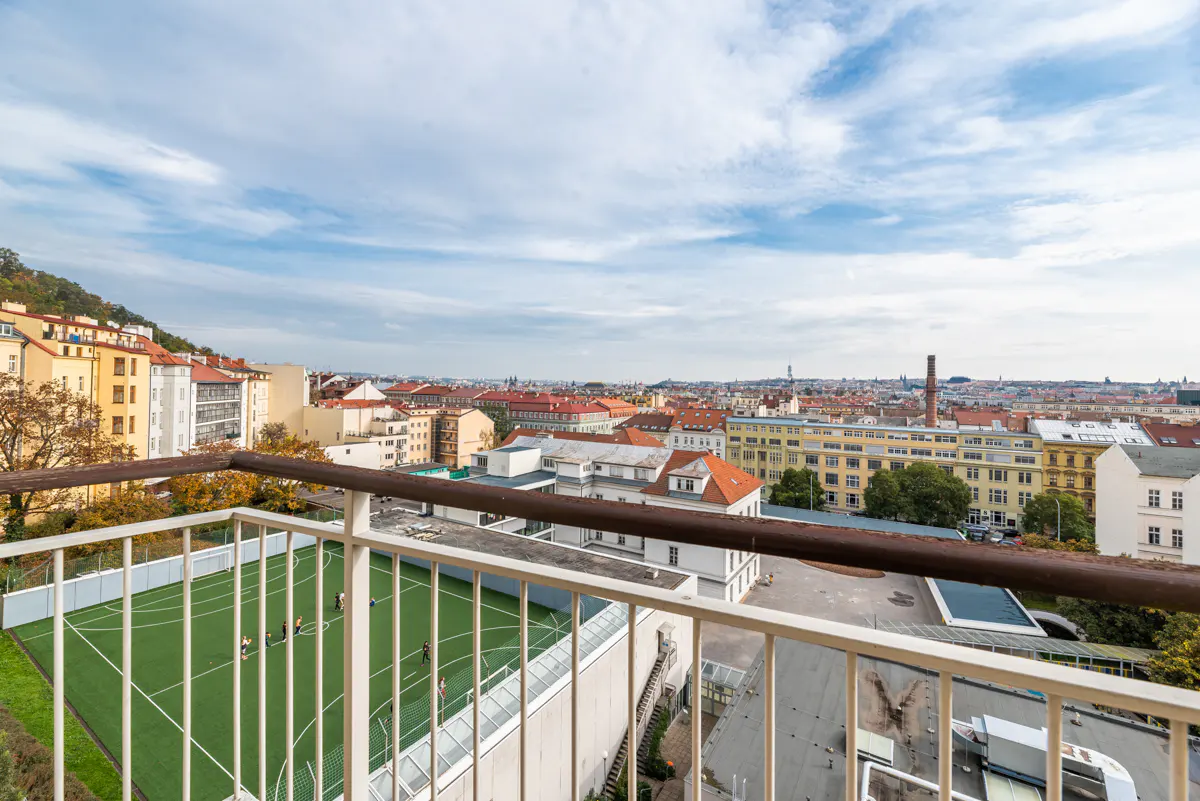 View from balcony overlooking a green soccer field with players, and a cityscape under a cloudy sky.