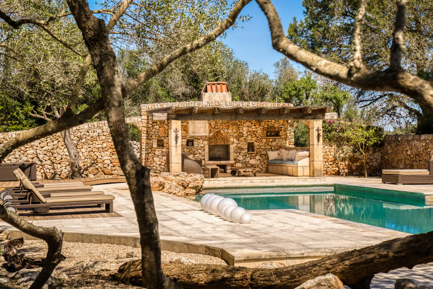 Outdoor pool area with stone cabana, fireplace, and lounge seating. Turquoise water reflects the blue sky. Trees frame the scene.