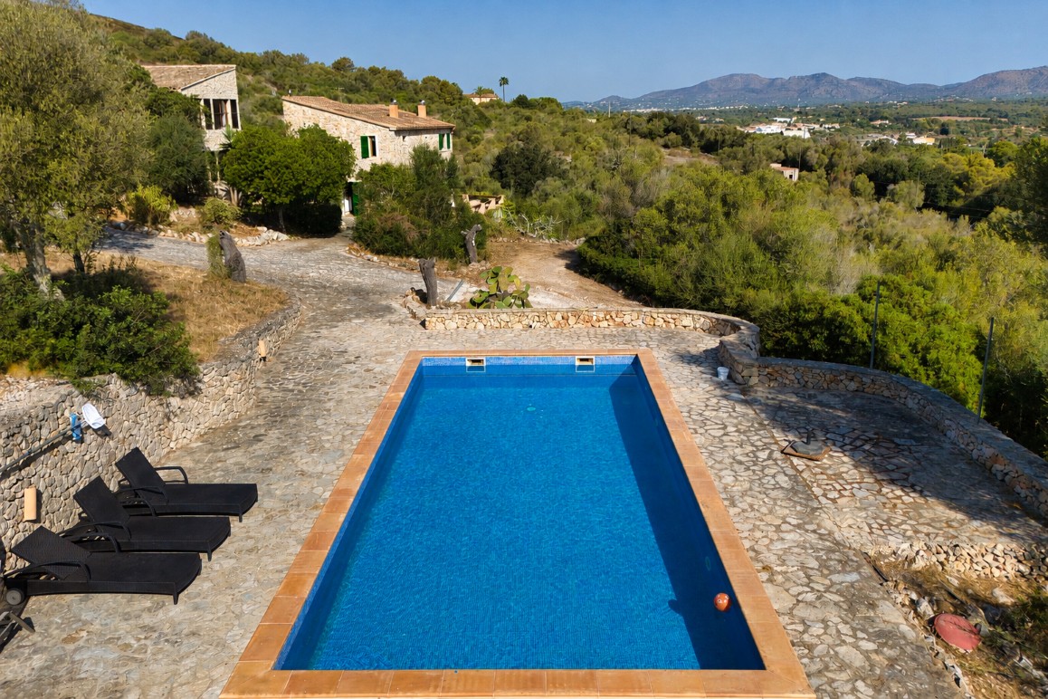Rectangular blue swimming pool on a stone patio with black lounge chairs, overlooking a rustic villa and hillside landscape. [AI-generated text]