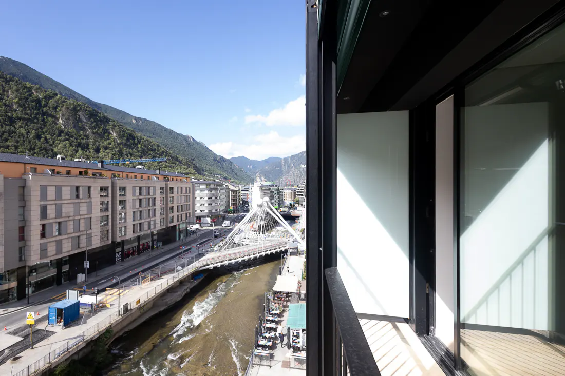 View from a balcony overlooking a city street, a river, a modern white bridge, and mountains under a clear blue sky.
