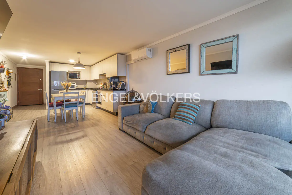 Open-plan living area with a grey sectional sofa, dining table, and modern kitchen with white cabinets and wood flooring.