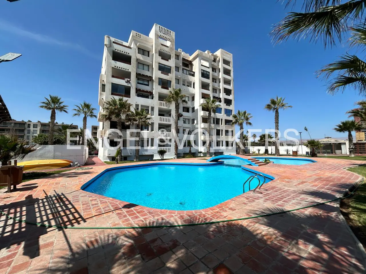 Exterior view of a white condo building with a pool and palm trees on a sunny day.