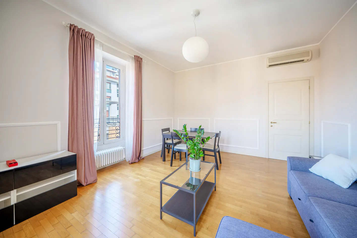 Bright living room with wood floors, blue sofa, and dining table. Pink curtains frame a window with a city view. A plant sits on a glass coffee table.