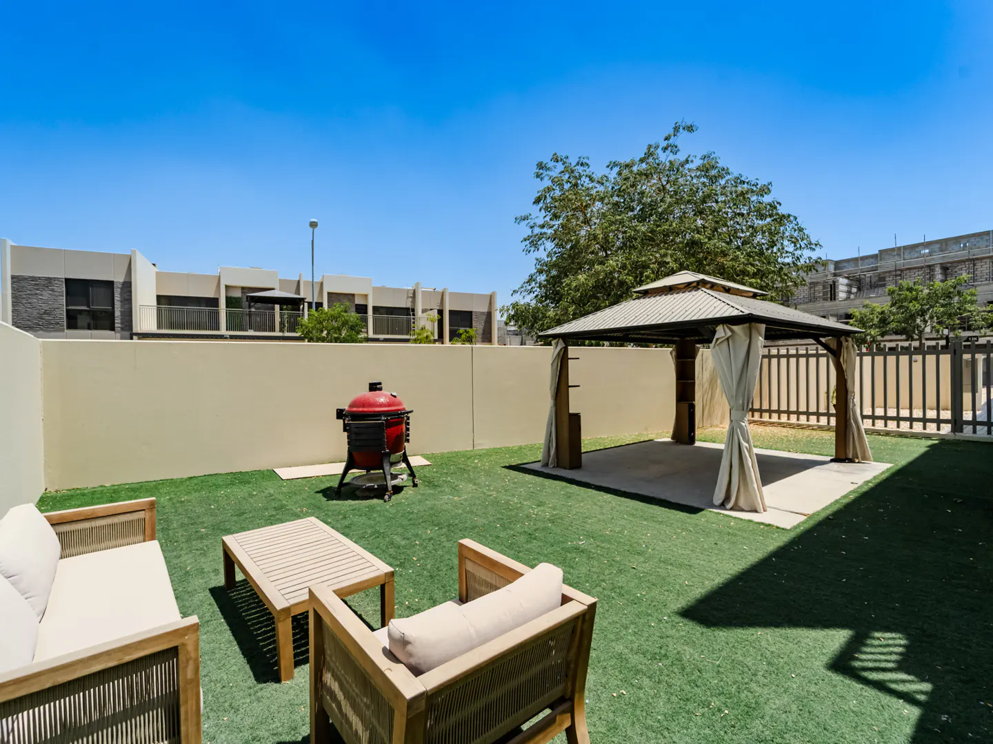 Backyard with artificial grass, a gazebo with curtains, a red charcoal grill, and outdoor lounge furniture.