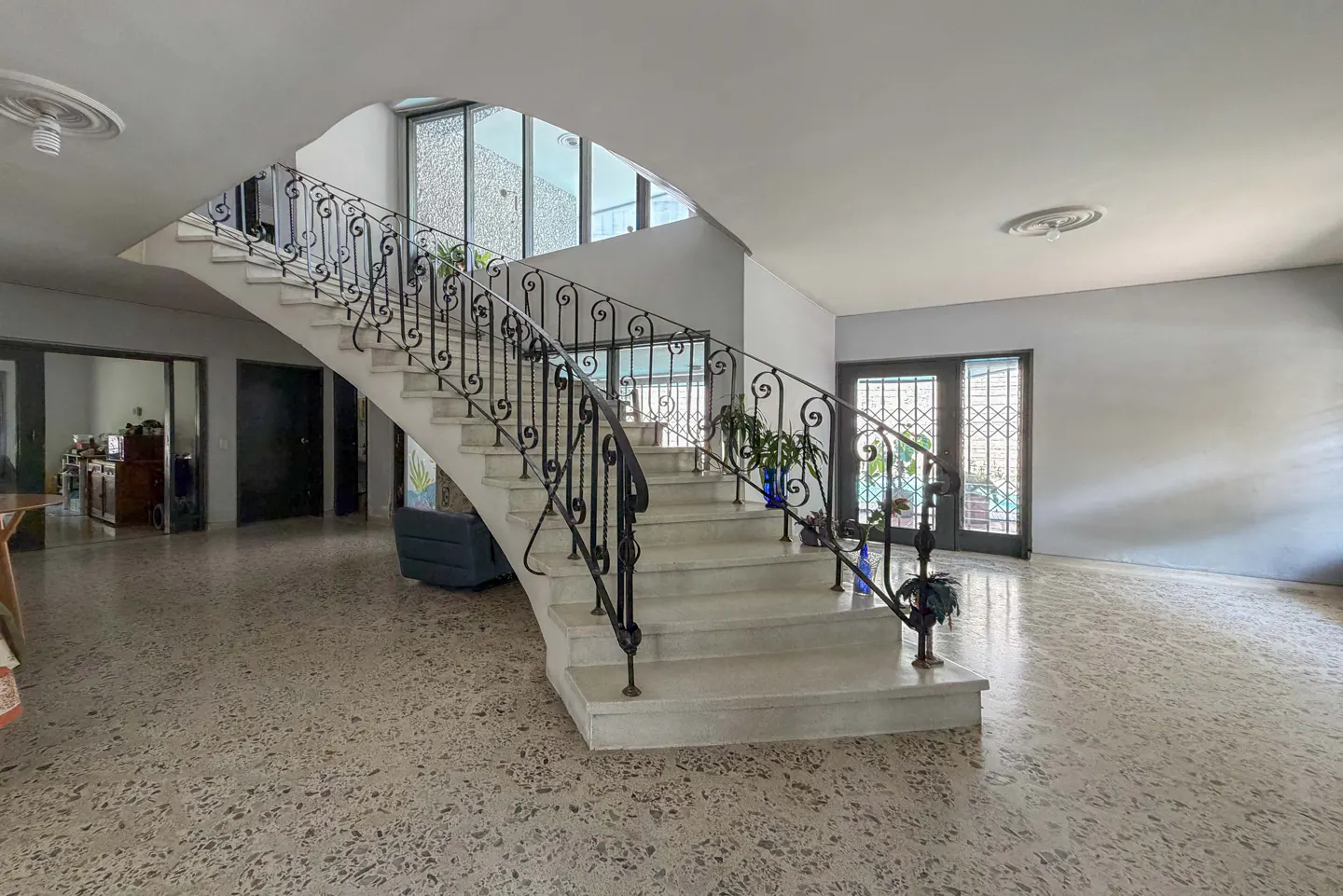 A bright foyer with a white staircase and black wrought iron railing. Potted plants decorate the stairs. Terrazzo flooring.