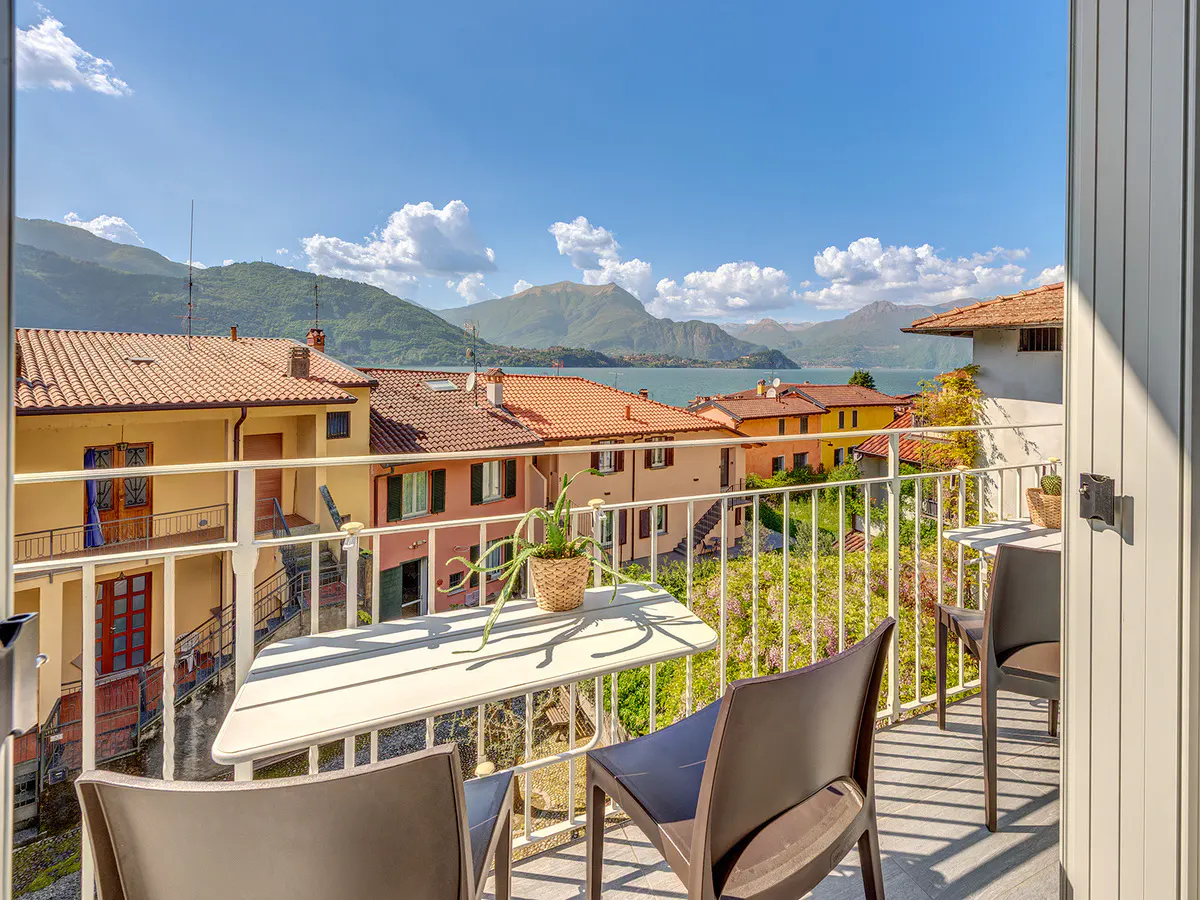Balcony view with table, chairs, and potted plant overlooking colorful buildings, lake, and mountains under a blue sky.