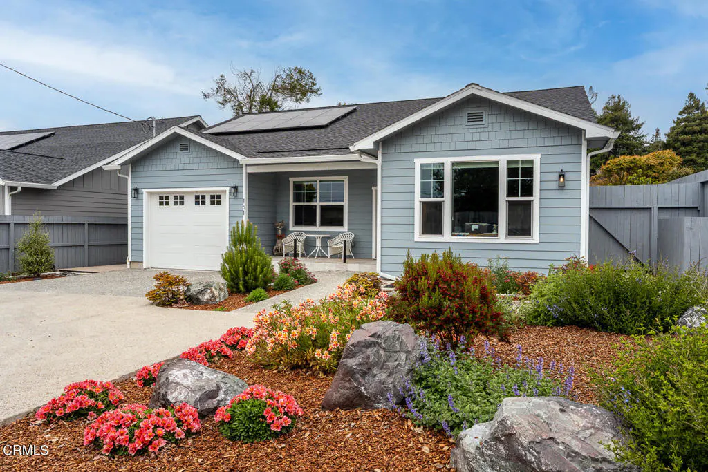 Single-story blue house with white trim, solar panels, a garage, and a landscaped front yard with rocks and flowers.