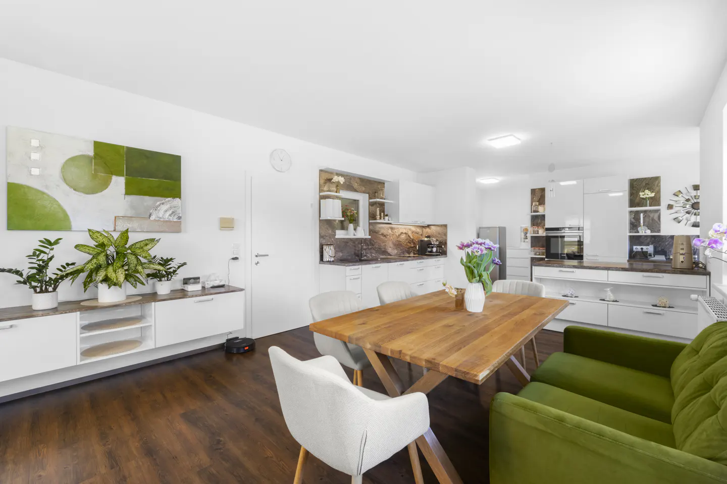 Bright, open-concept living space with a wooden dining table, green sofa, and white kitchen cabinets. Artwork and plants add color.