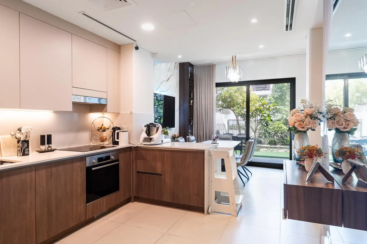 A bright, modern kitchen with wood cabinets, white countertops, and stainless steel appliances. A dining area with a garden view is visible.