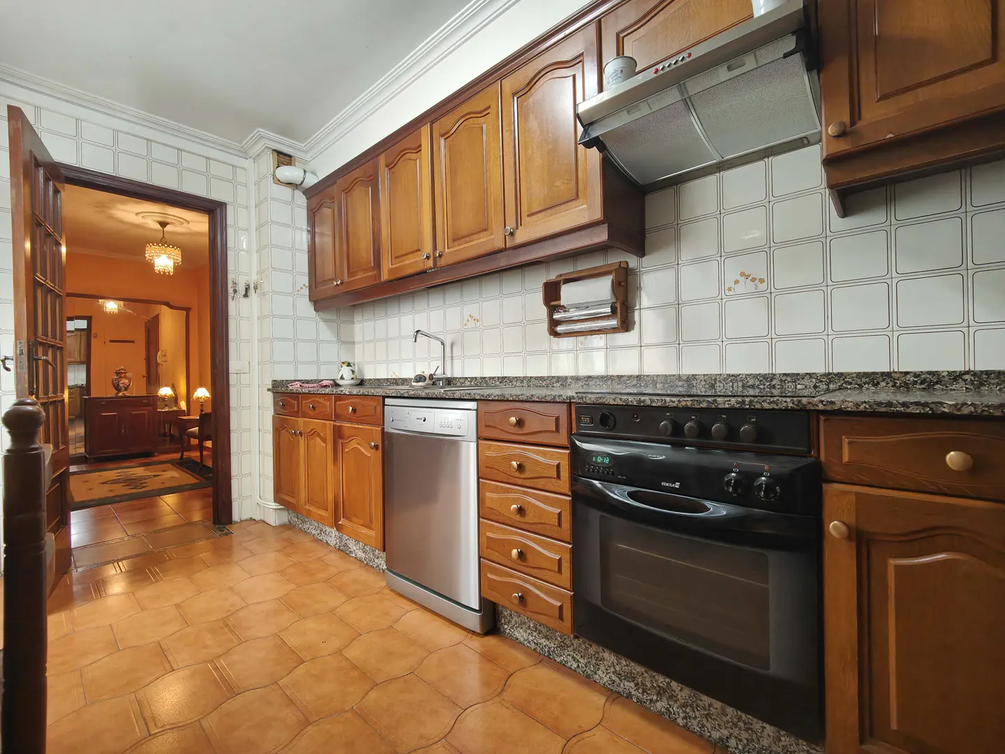 A kitchen with brown cabinets, a black oven, and a stainless steel dishwasher. The floor is tiled in brown, and an open doorway leads to a living room.