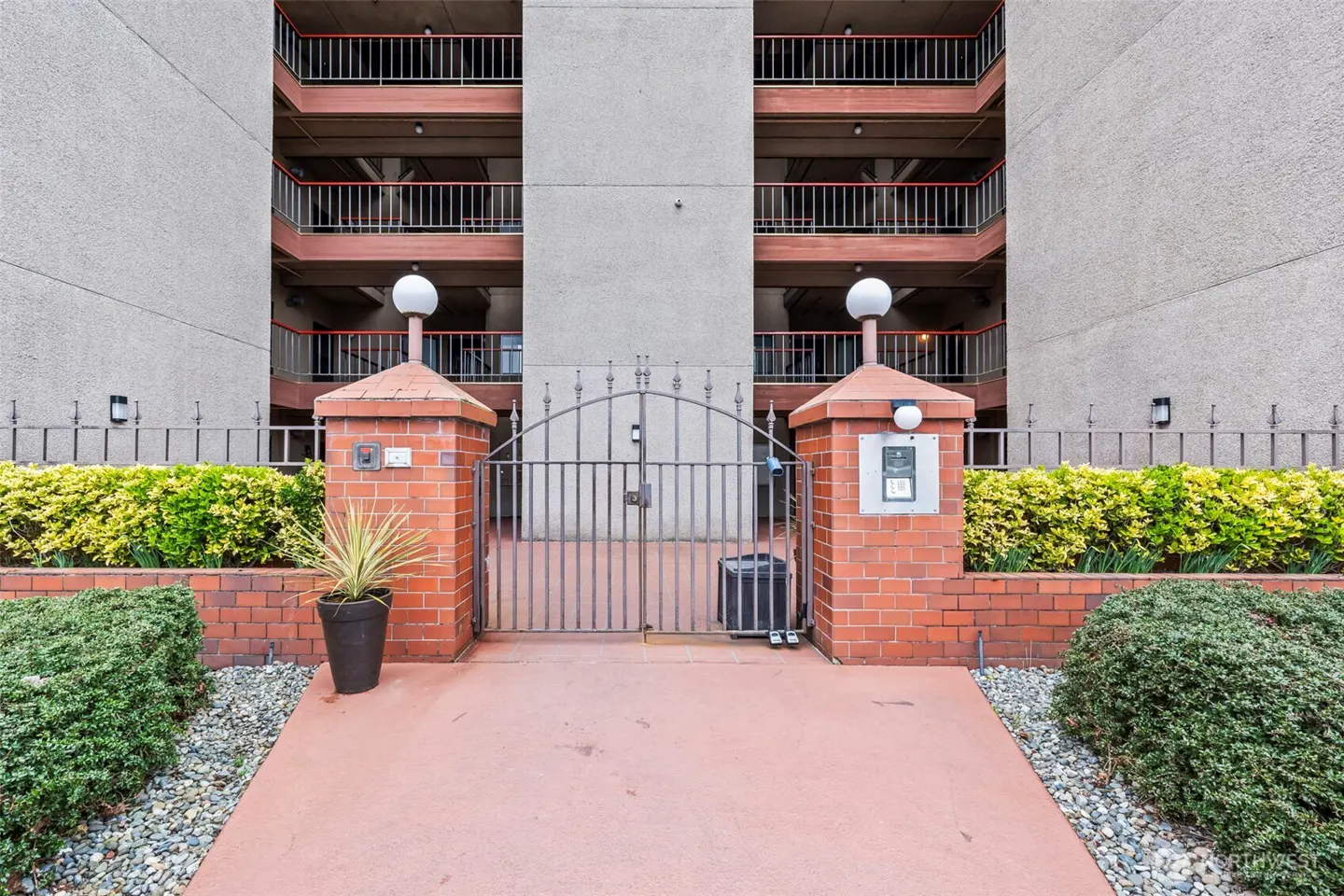 A gated entrance to a multi-story apartment building with brick pillars, a metal gate, and manicured landscaping.