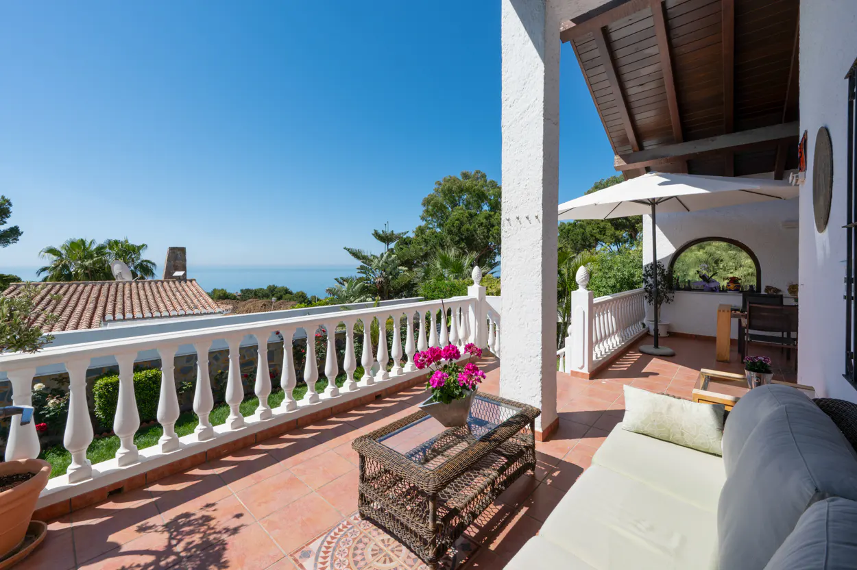 Outdoor patio with white balustrade, terracotta tiles, and wicker furniture. Ocean view in the background under a clear blue sky.