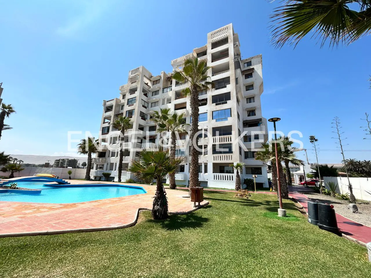 Exterior view of a white condo building with a pool, palm trees, and green lawn under a blue sky.