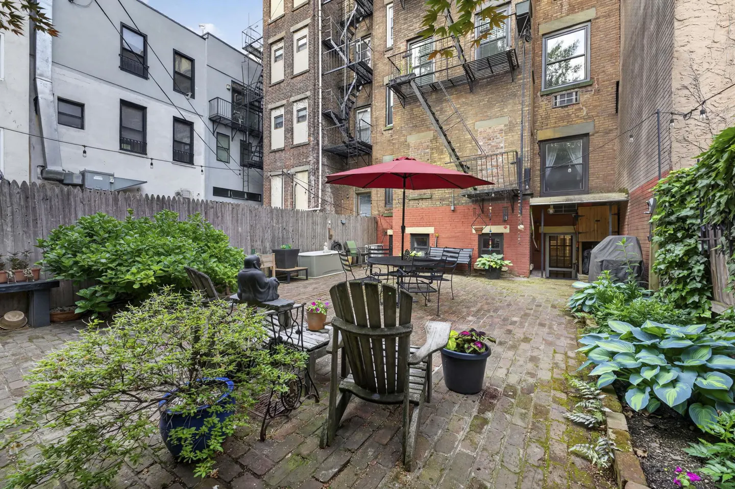A brick patio features a red umbrella, outdoor seating, and lush potted plants against a backdrop of urban brick buildings.