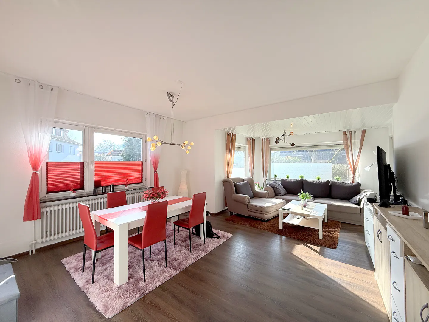 Open-concept living and dining area with wood floors, white walls, and red accents. A white table with red chairs sits on a pink rug.