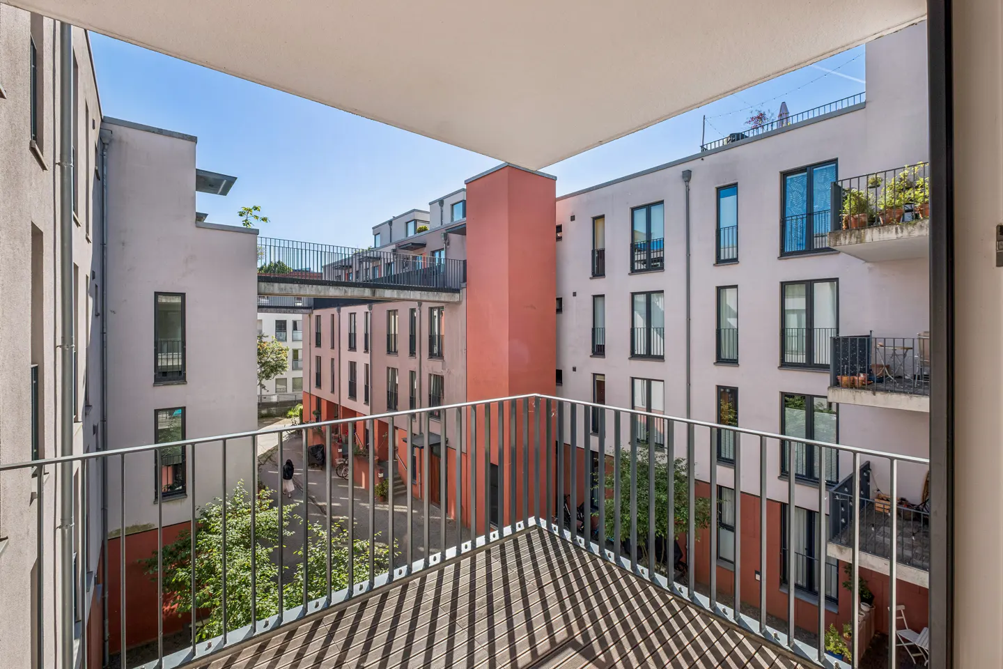 View from a balcony overlooking a modern apartment courtyard with light-colored buildings, red accents, and a footbridge.