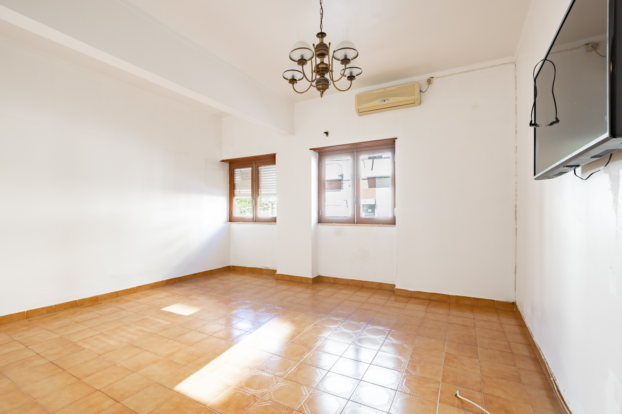 Bright, empty room with white walls, brown tile floor, two windows, chandelier, AC unit, and a TV mounted on the wall.