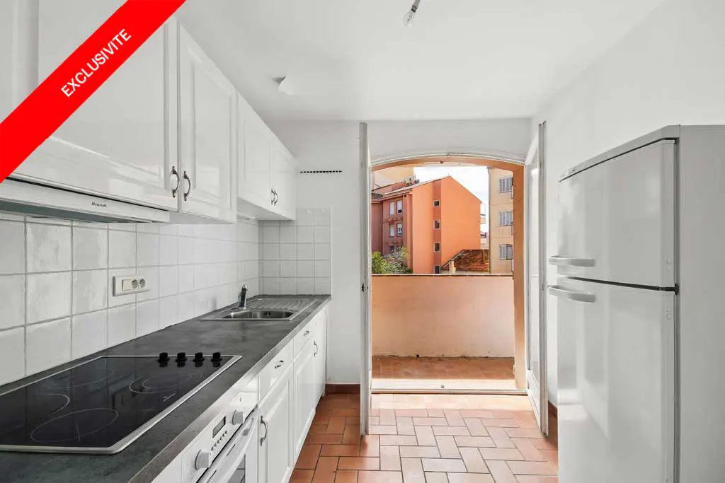 A bright kitchen with white cabinets, a black countertop, and a view to a balcony. A red banner reads "Exclusivite".