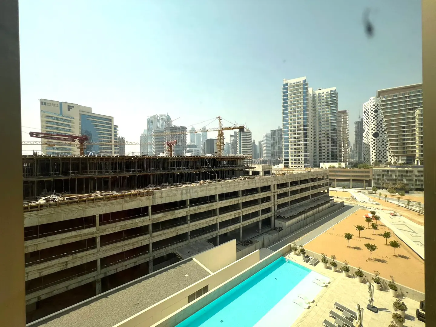 View from a window showing a bright blue swimming pool, a sandy courtyard, and a large construction site with city towers.