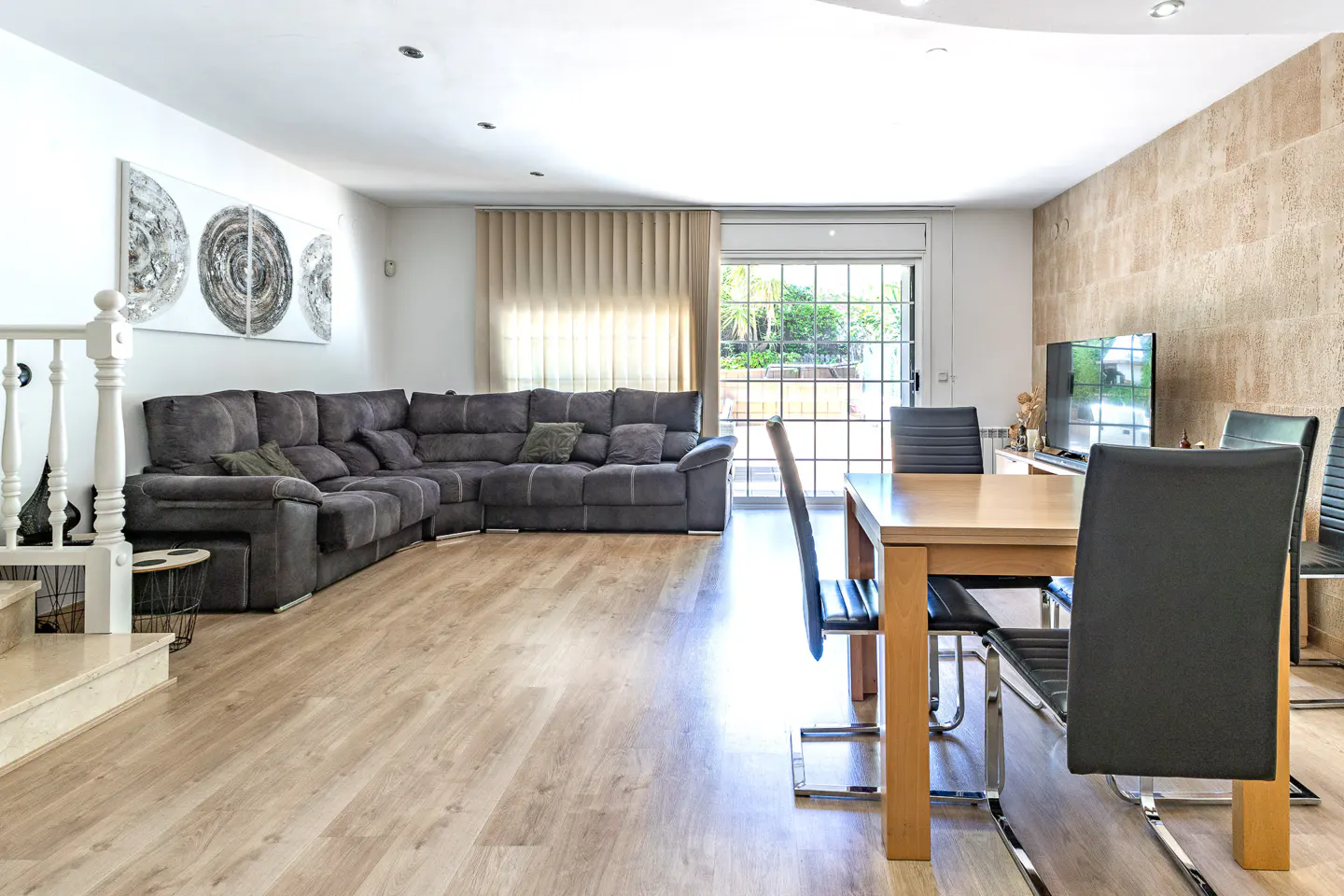 Living room with a gray sectional sofa, wood floors, and a dining table with black chairs. Artwork on the wall.