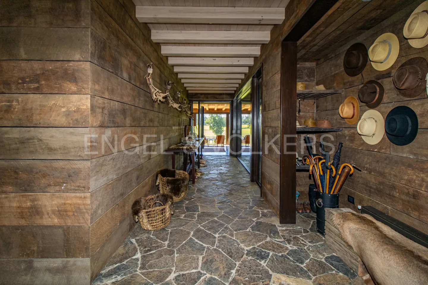 Rustic hallway with stone floor, wood walls, and white ceiling beams. Hats and umbrellas are displayed on the right wall. Baskets line the left.
