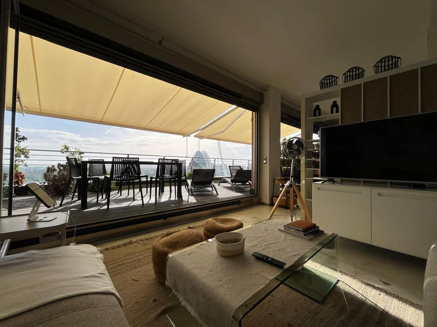 Modern living room with a glass coffee table, TV unit, and large sliding doors opening to a sunny balcony with dining set.