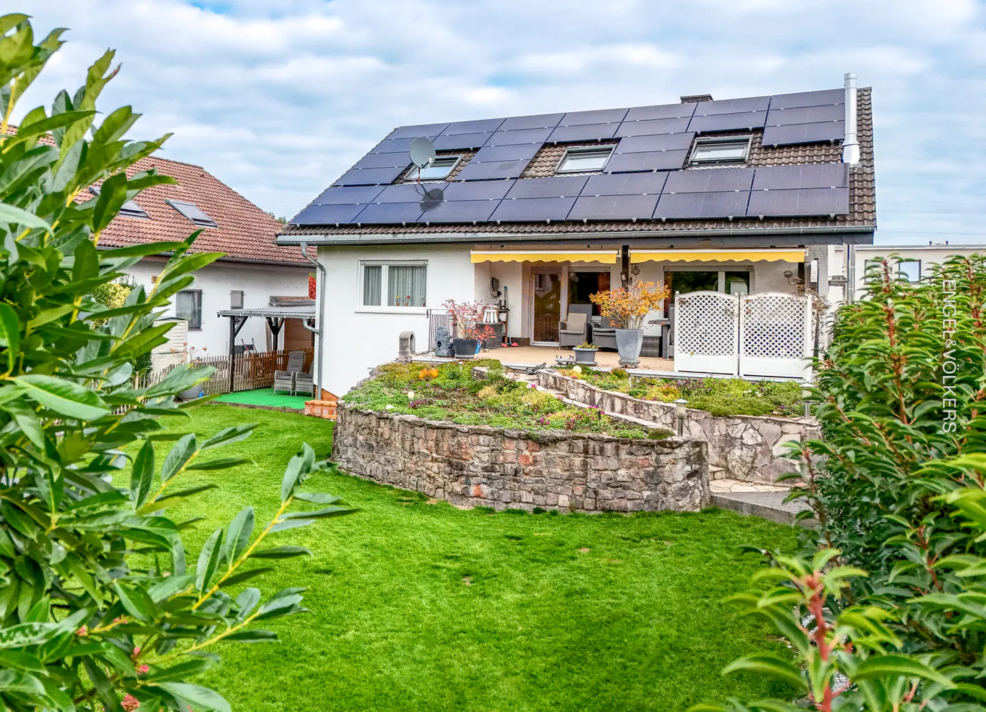 A white house with solar panels on the roof, a stone wall, and a green lawn.
