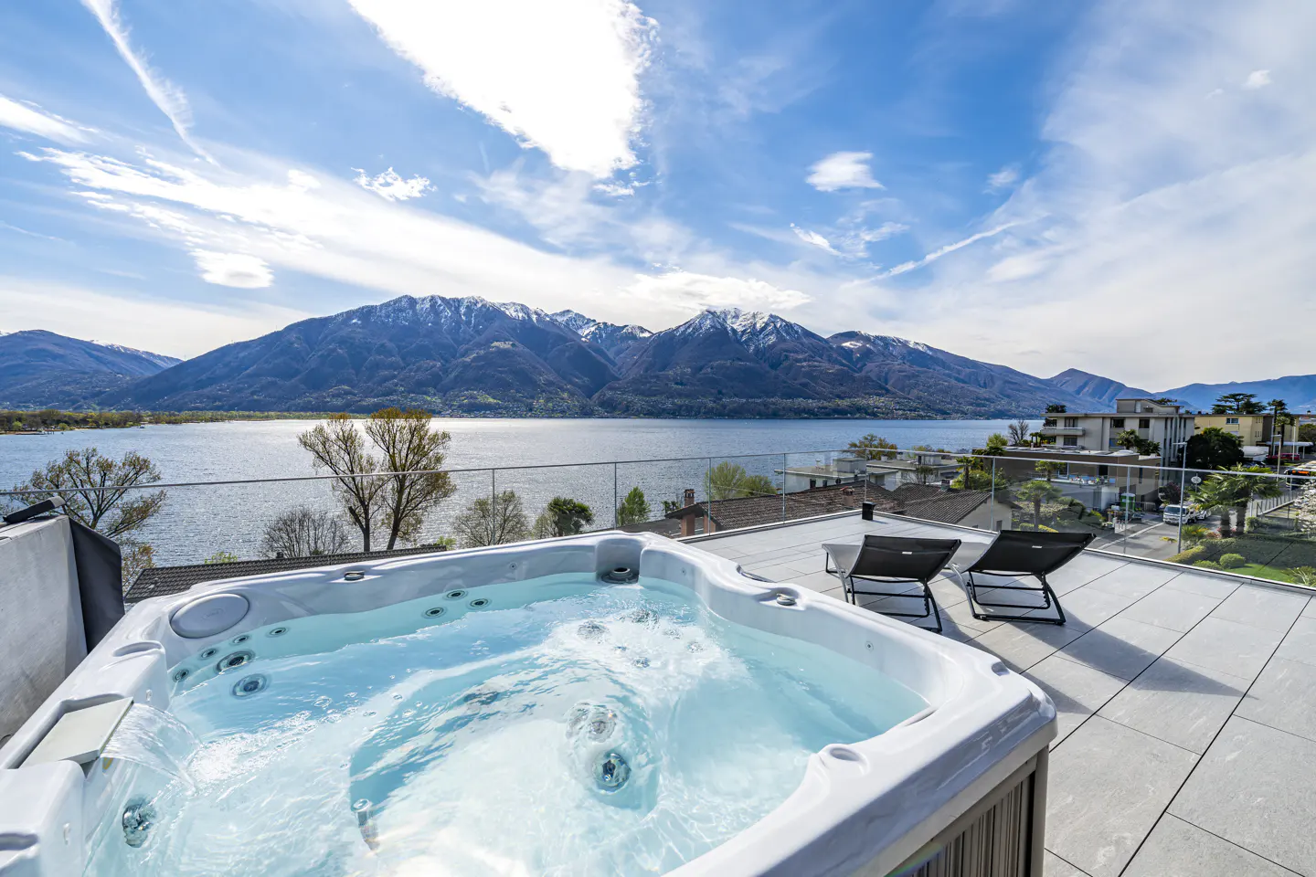 Rooftop patio with a hot tub, two lounge chairs, and a scenic view of a lake and mountains under a blue sky.