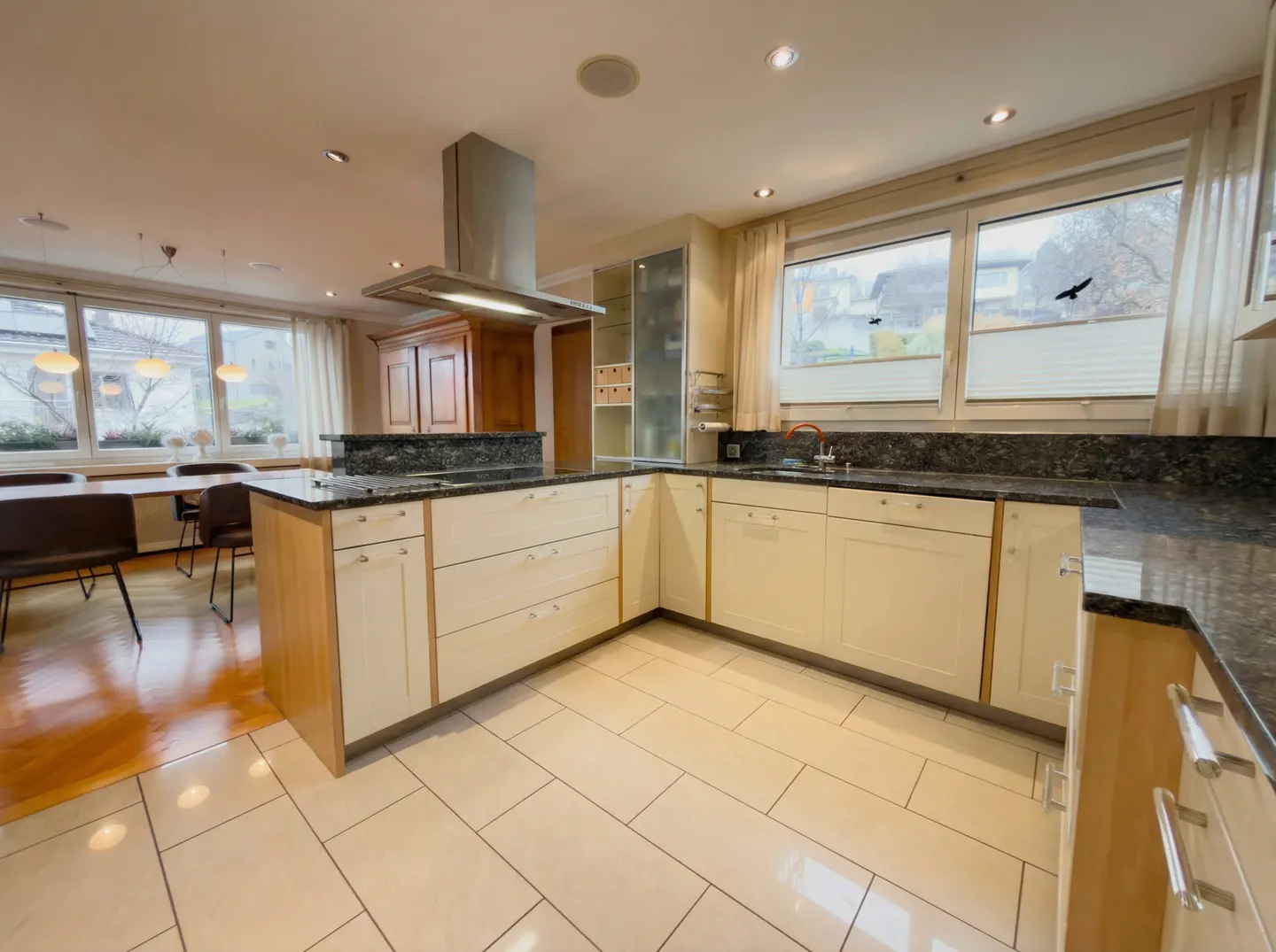 A bright kitchen with cream cabinets, granite countertops, and stainless steel appliances. A dining area is visible in the background.