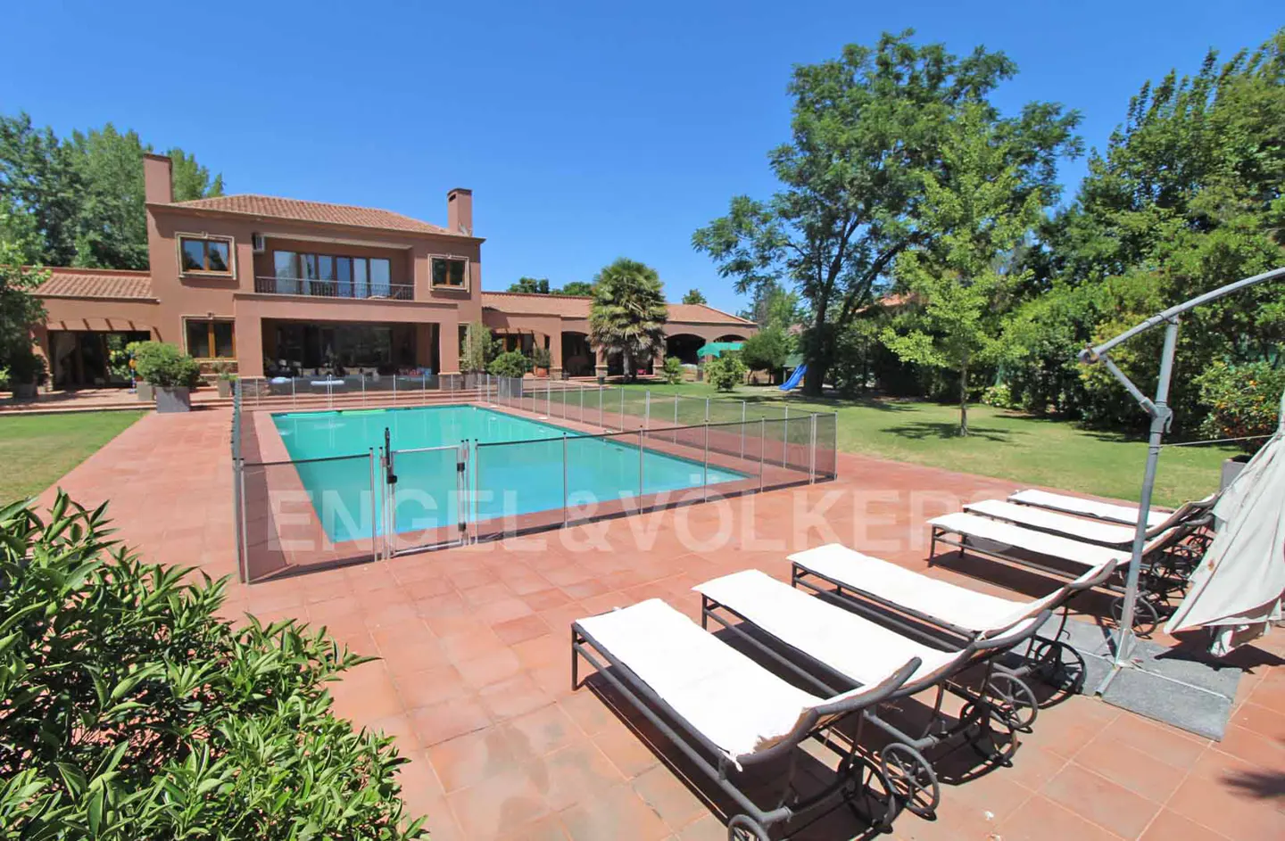 Exterior view of a two-story terracotta house with a pool, lounge chairs, and green trees under a blue sky.