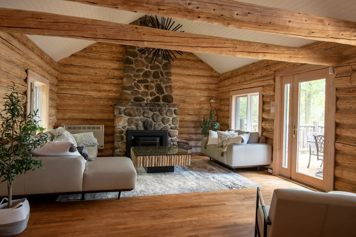 Living room in a log cabin with a stone fireplace, light gray sofas, wood floors, and a door to a deck.