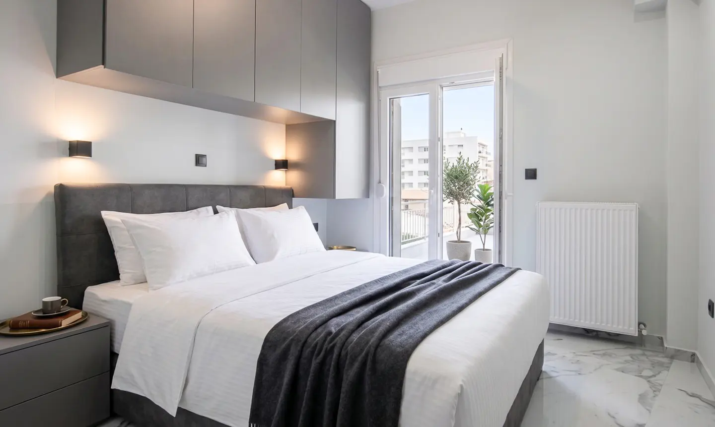 A modern bedroom with a gray headboard, white linens, and a gray throw blanket. A balcony with plants is visible through the open doors.