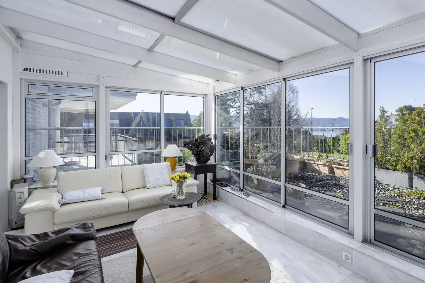 Bright sunroom with white walls, glass ceiling, and large windows overlooking a garden and distant water. Sofas and a wooden table furnish the space.