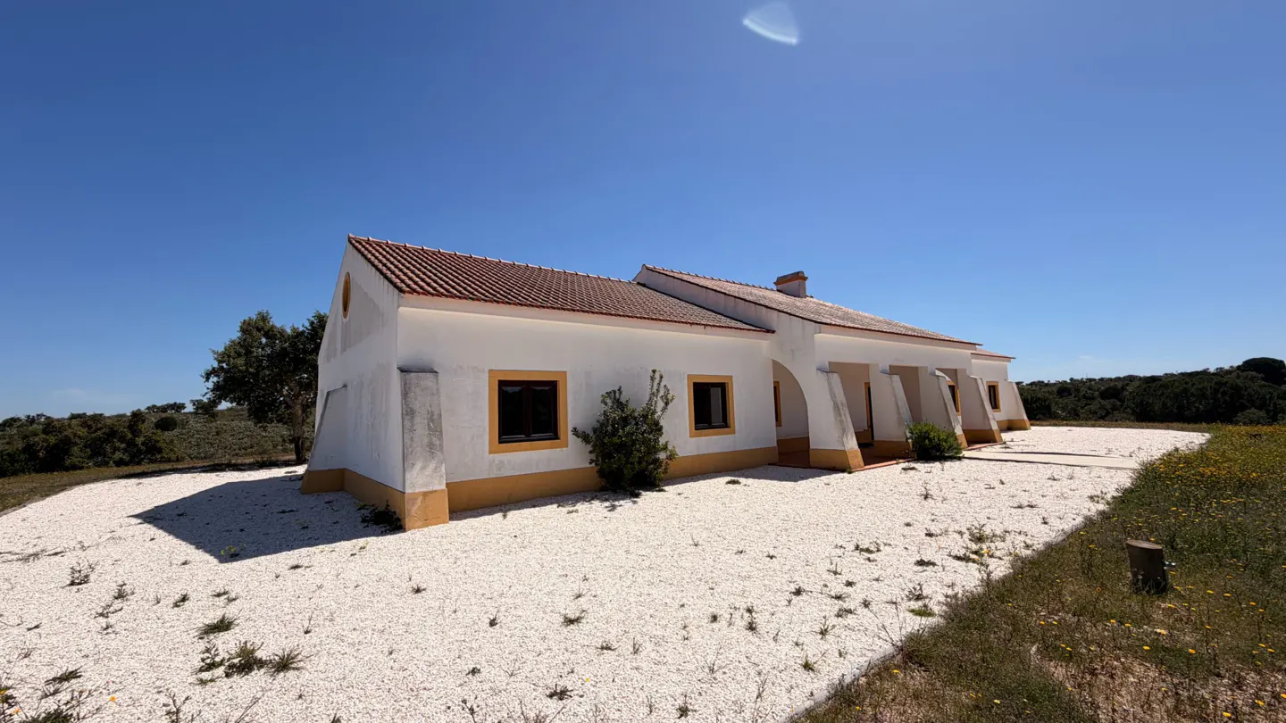 A white, single-story house with a terracotta roof and yellow trim, set on a gravel yard under a clear blue sky.