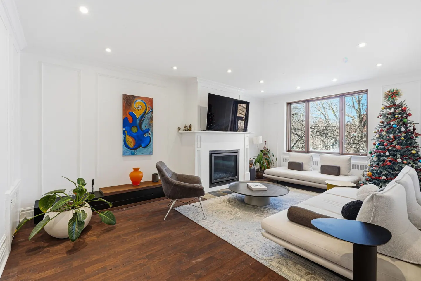 Bright living room with hardwood floors, white walls, and recessed lighting. A fireplace, sofas, and a decorated Christmas tree are also visible.