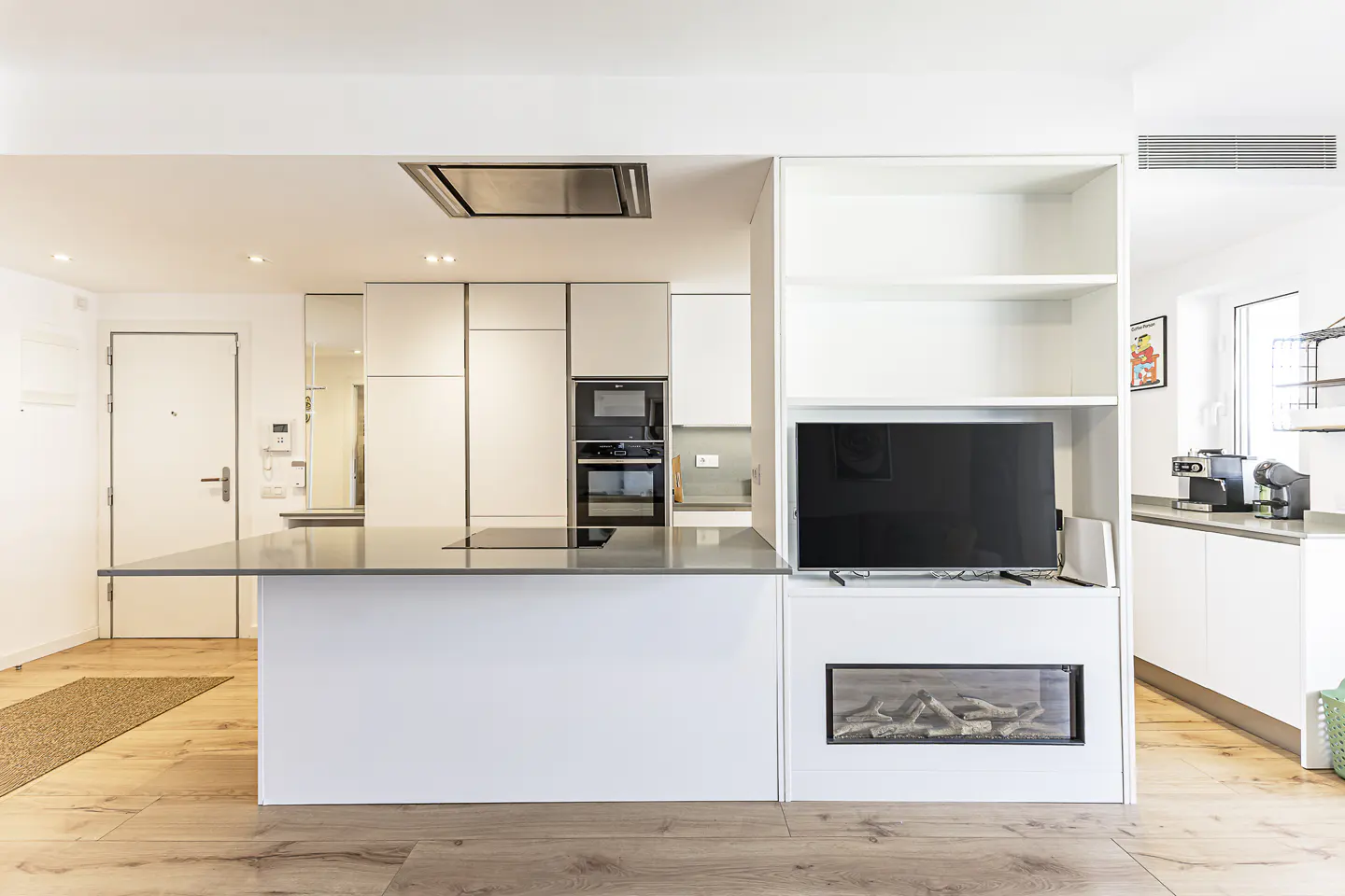 Modern kitchen with white cabinets, a gray island, and wood floors. A TV and fireplace are built into a shelving unit.