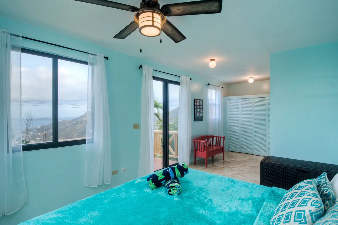 A bright bedroom with aqua walls, a ceiling fan, and a view. A turquoise bedspread and pillows add color. A window and sliding glass door let in natural light.
