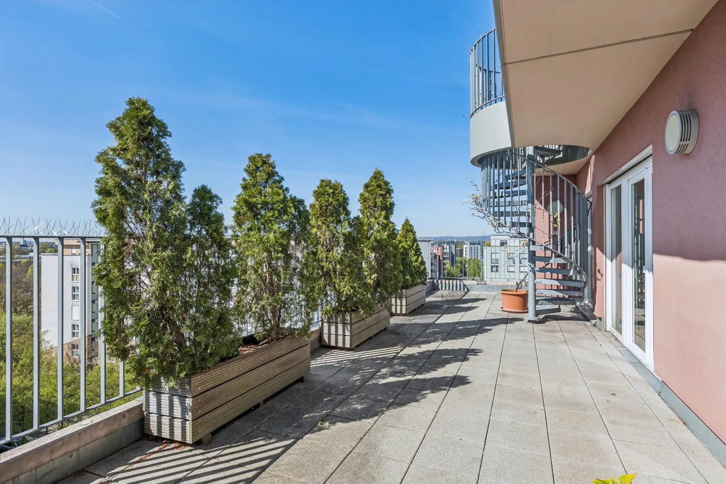 A rooftop patio with potted evergreen trees, a spiral staircase, and a view of the city on a sunny day.