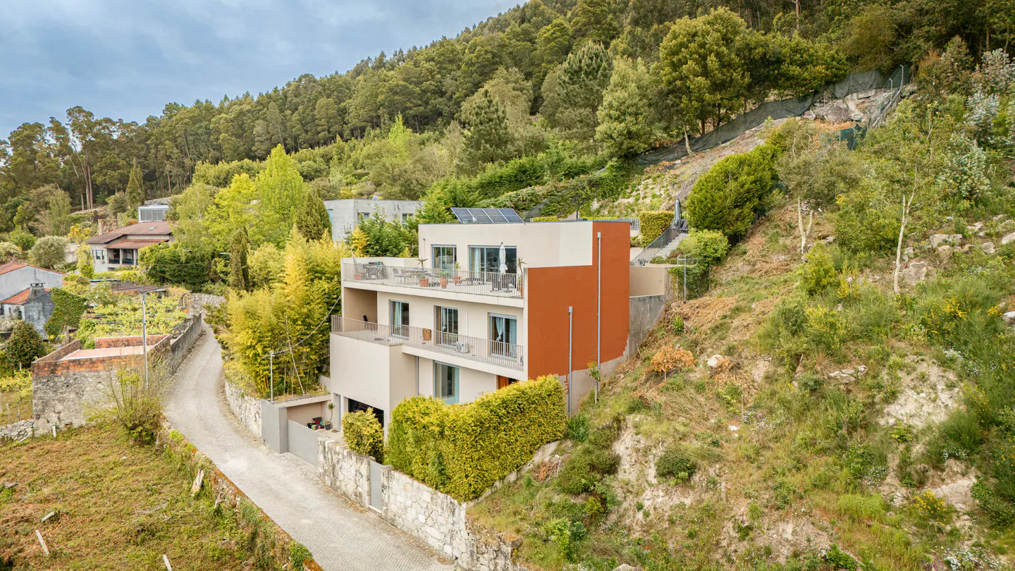 A modern three-story house with balconies, beige walls, and a rust-colored accent wall, nestled on a green hillside.