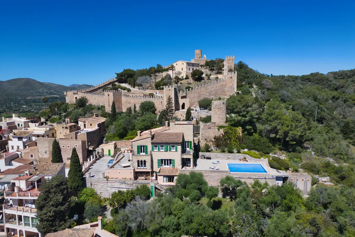 Aerial view of Capdepera Castle, Mallorca, Spain, atop a hill overlooking a town, with a blue pool in the foreground.