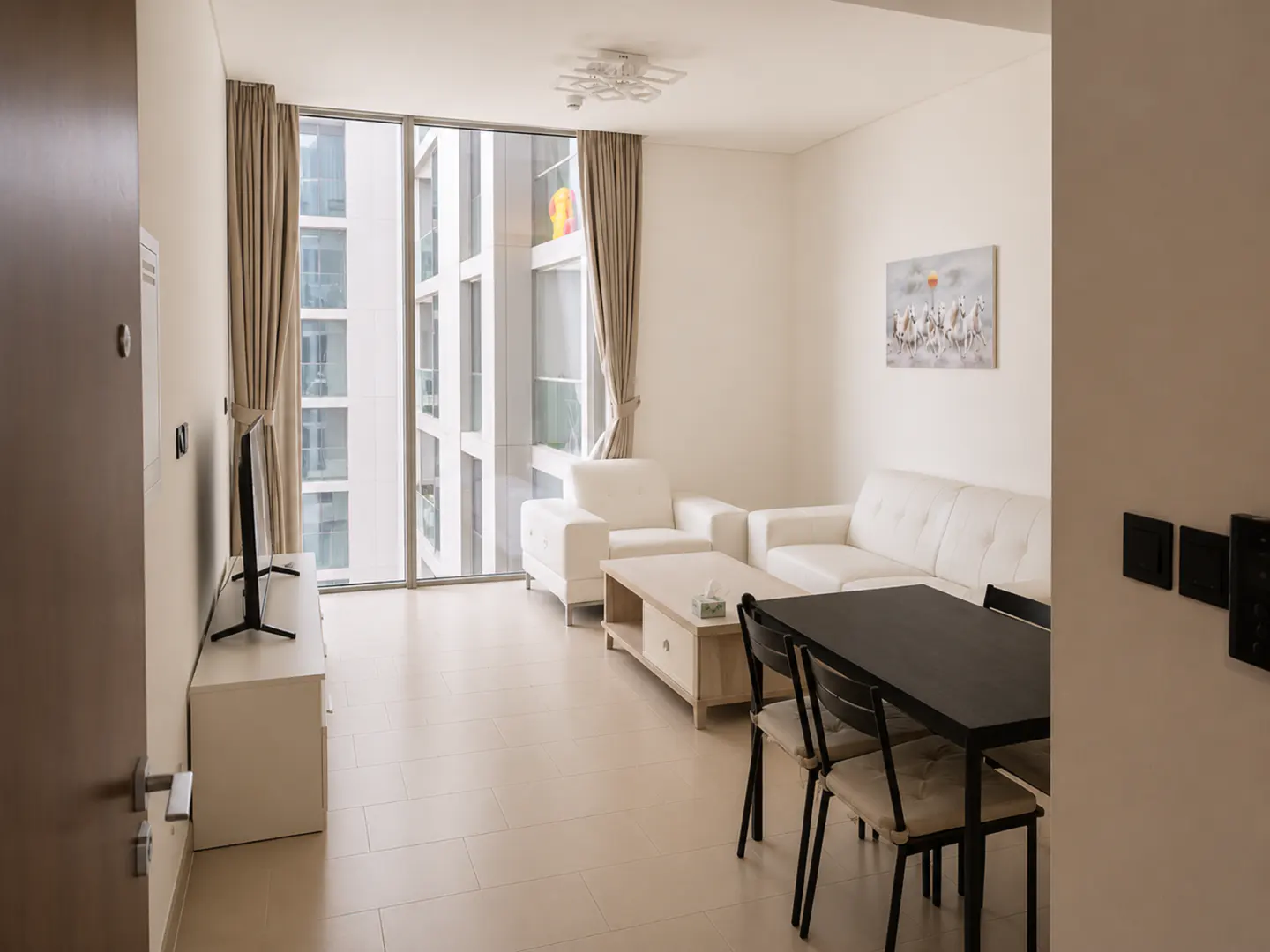 Bright living room with white walls, furniture, and tile floors. A black dining table sits near a white sofa and chairs. Large windows offer a city view.