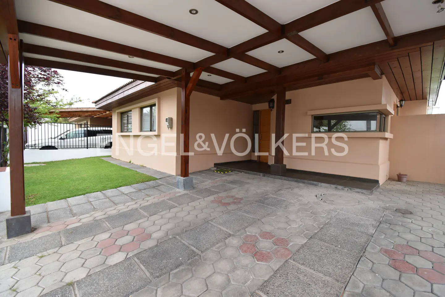 Exterior view of a beige house with a brown wood carport and a patterned stone driveway. Green lawn visible.