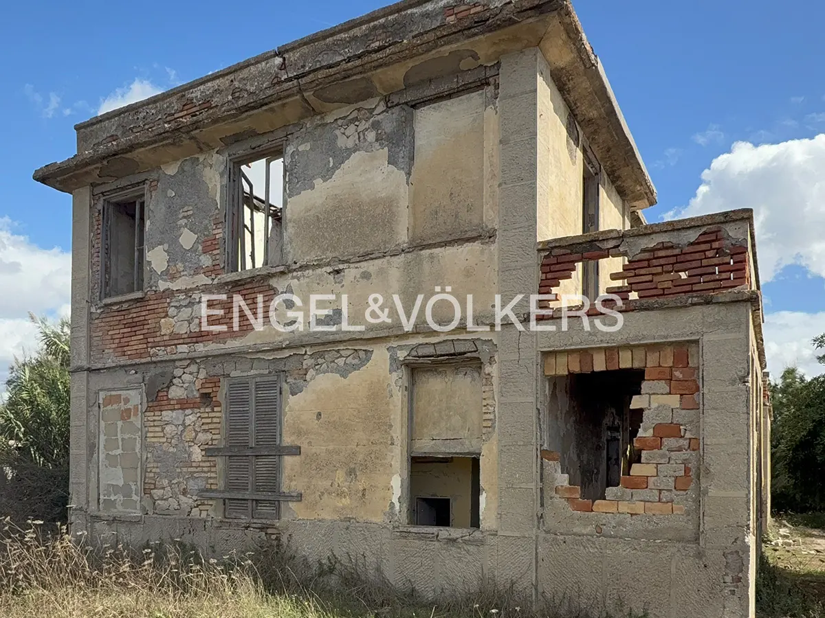 A dilapidated two-story house with crumbling beige stucco, exposed red brick, and empty window frames under a blue sky.